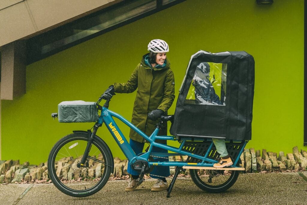 Parent in waterproof rain jacket riding cargo bike with child protected in Eco Pop Top